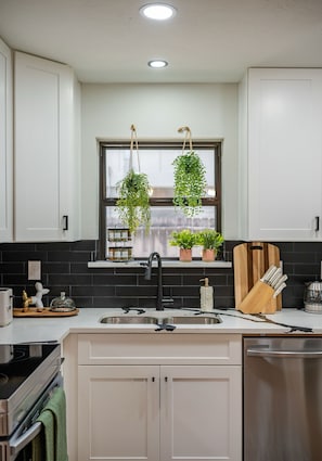 A kitchen with white cabinets, a black backsplash, a window with hanging plants, a sink, and a stove. - Cozy 5-bedroom house in adorable Spring with pool (Spring)