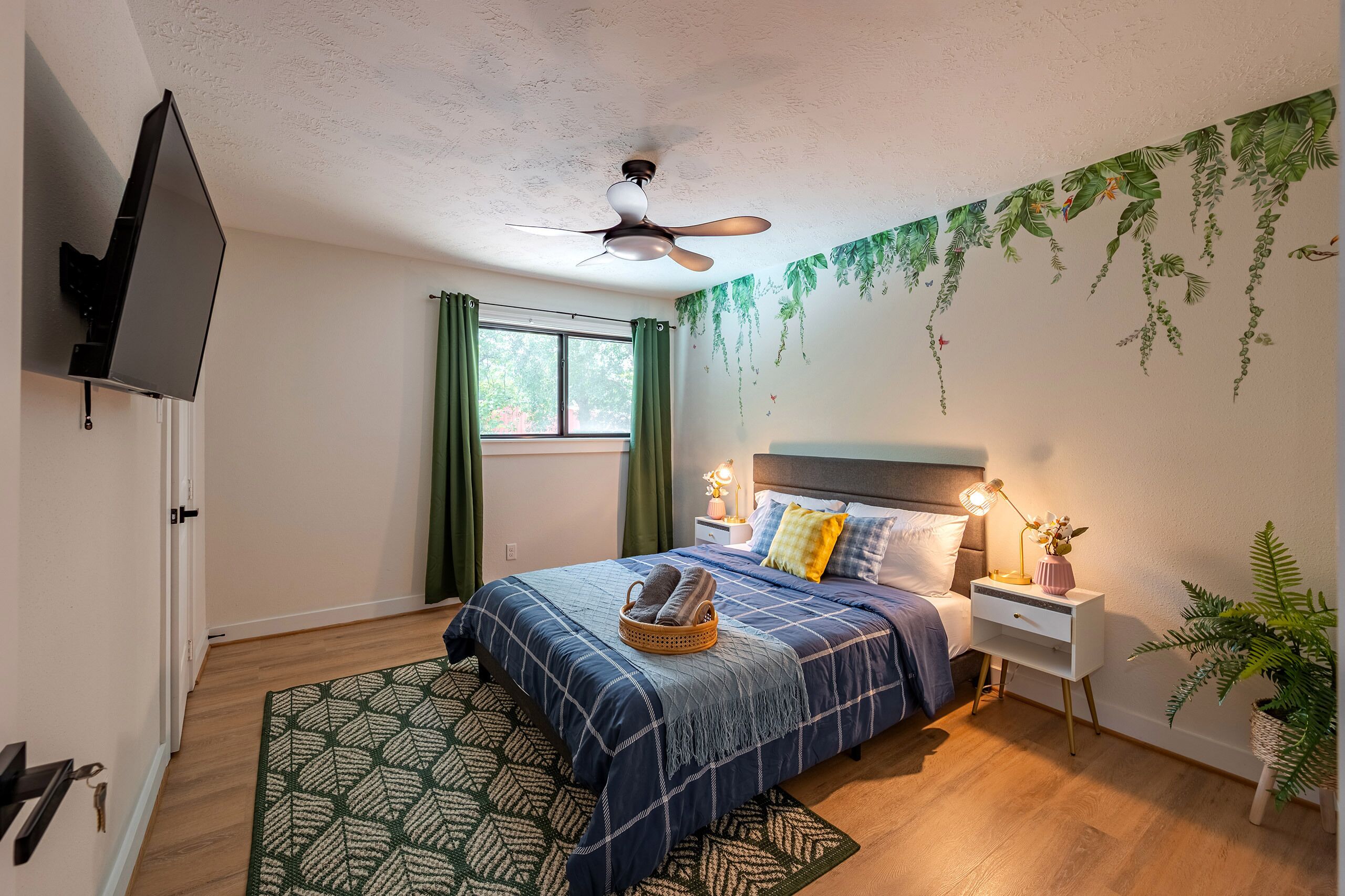 Ein Schlafzimmer mit Bett, Nachttisch, Lampe, Deckenventilator, Fenster mit Vorhängen und einer Wand mit hängenden Pflanzen.