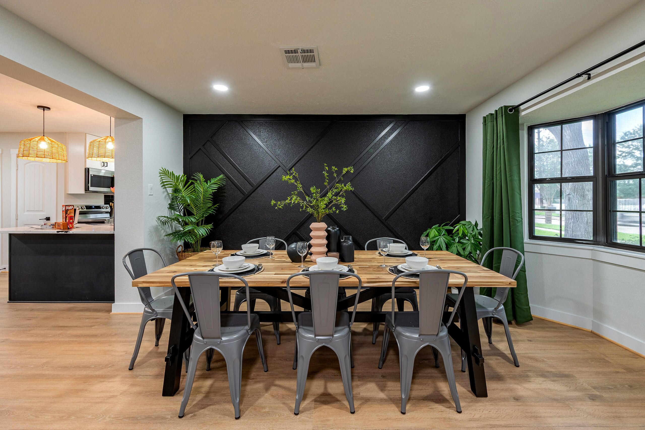 A dining area with a wooden table set for six, surrounded by grey chairs, with a black textured wall behind and a window with green curtains to the right.