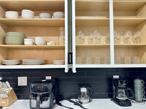 A kitchen with wooden shelves holding plates and glasses, a coffee maker, and a kettle.