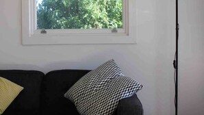 Una habitación con un sofá negro, un cojín decorativo estampado, una lámpara de pie y una ventana con vistas a árboles.
