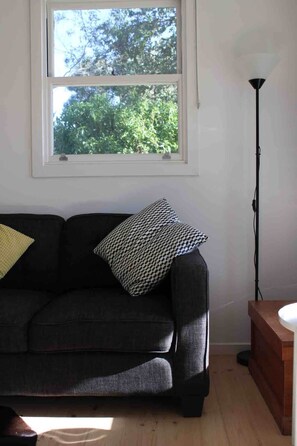 Ein Zimmer mit einer schwarzen Couch, einem gemusterten Kissen, einer Stehlampe und einem Fenster mit Blick auf Bäume.