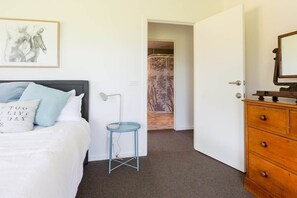 Une chambre à coucher comprenant un lit, une table de chevet, une commode, une porte donnant sur une salle de bain et un tableau encadré accroché au mur.