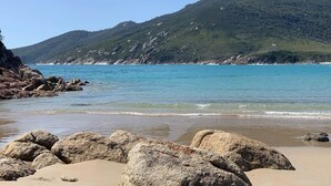 Ein Strand mit großen Felsen im Vordergrund, blauem, klarem Meer und einer bewachsenen Hügelkulisse im Hintergrund.