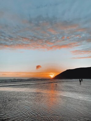 Eine Strandansicht mit Sonnenuntergang, ruhigem Wasser und der Silhouette einer Hügelkette.