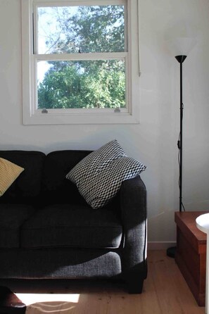 Ein Zimmer mit einem dunkelgrauen Sofa, einem gemusterten Kissen, einer Stehlampe und einem Fenster mit Blick auf Bäume.