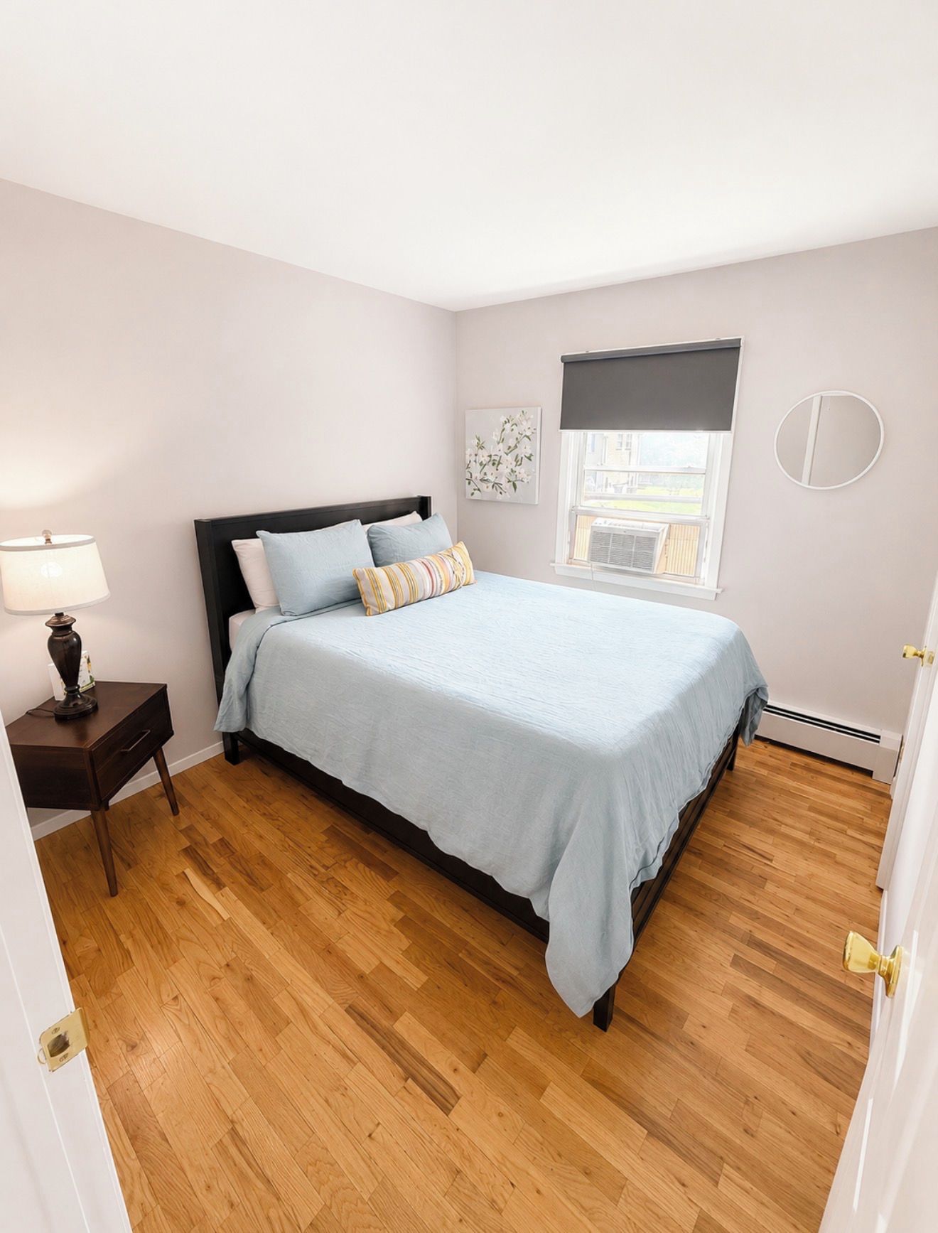 Un dormitorio con cama, mesita de noche, lámpara, ventana con persianas y espejo montado en la pared.