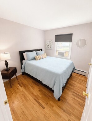 Un dormitorio con cama, mesita de noche, lámpara, ventana con persianas y espejo montado en la pared.