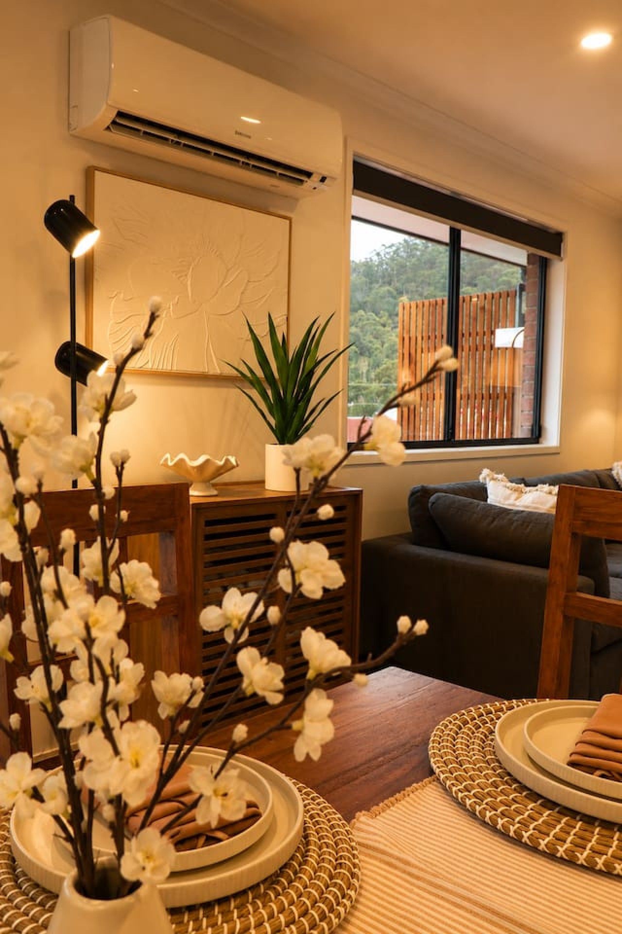A modern living room with a sofa, a dining table with plates, a potted plant, and a wall-mounted air conditioner.