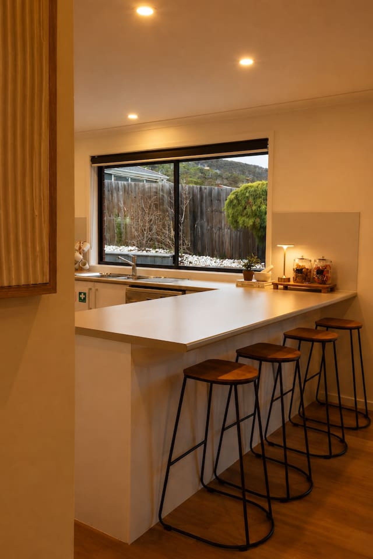 A modern kitchen with a large window, bar stools, and a view of a backyard.