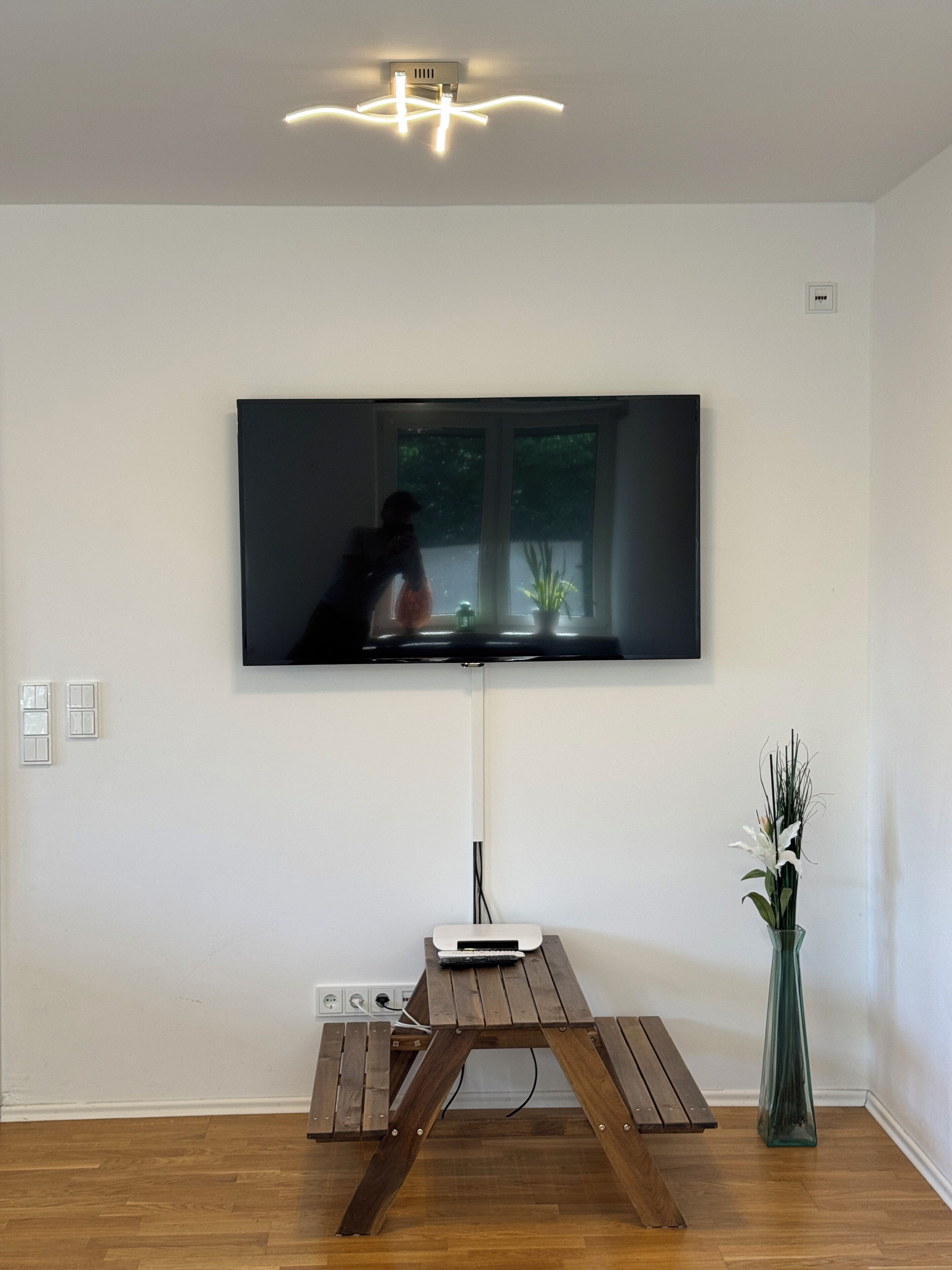 A flat-screen TV mounted on a wall above a wooden table with a vase of flowers.