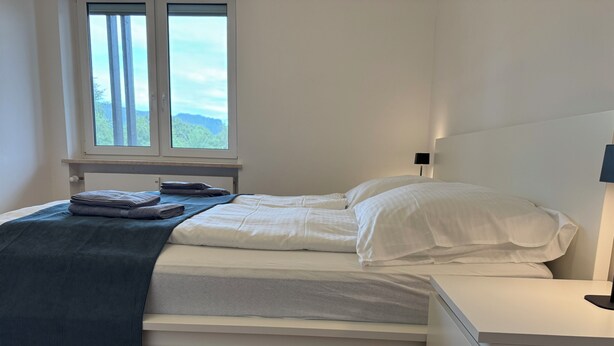 Ein ordentlich bezogenes Bett mit weißen Leinen, einer gefalteten blauen Decke und einem Fenster mit Blick auf Grünflächen.