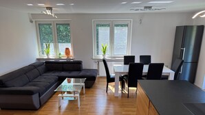 Ein modernes Wohnzimmer mit Ecksofa, einem Esstisch mit Stühlen, einer Küchenzeile mit Kühlschrank und großen Fenstern mit Pflanzen.