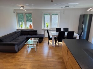 A modern living space with a sectional sofa, a dining table with chairs, a kitchen area with a refrigerator, and large windows with plants. - Bergblick Buchenhöhe: modern vacation rental at 1,000 m, surrounded by forest. (Berchtesgaden)