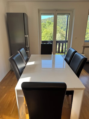 A dining area with a white table and black chairs, a black refrigerator, and a view of a balcony with a wooden railing. - Bergblick Buchenhöhe: modern vacation rental at 1,000 m, surrounded by forest. (Berchtesgaden)
