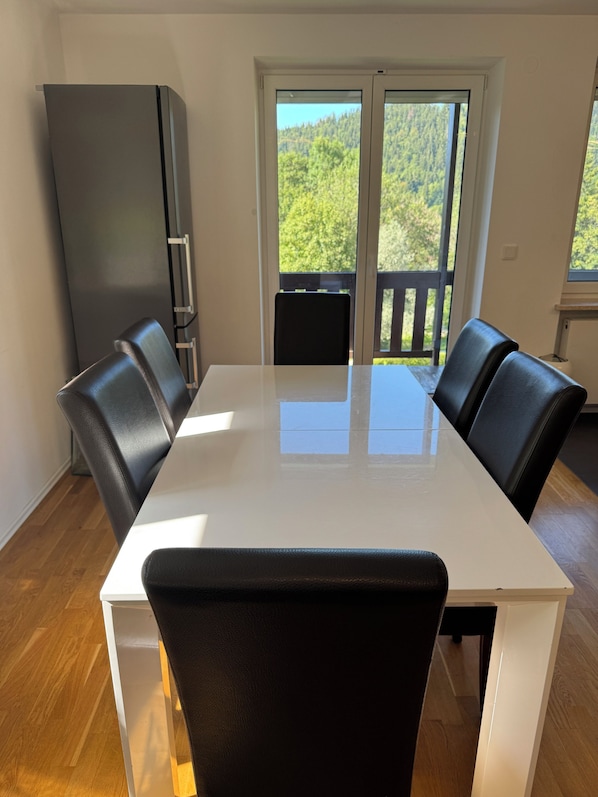 A dining area with a white table and black chairs, a black refrigerator, and a view of a balcony with a wooden railing.