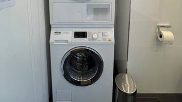 A stacked washer and dryer unit in a bathroom with a paper towel dispenser and a trash can.