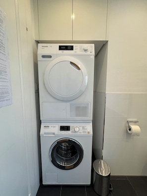 A stacked washer and dryer unit in a bathroom with a paper towel dispenser and a trash can.