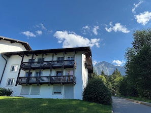 A two-story building with balconies, a road in the foreground, and mountains in the background. - Bergblick Buchenhöhe: modern vacation rental at 1,000 m, surrounded by forest. (Berchtesgaden)
