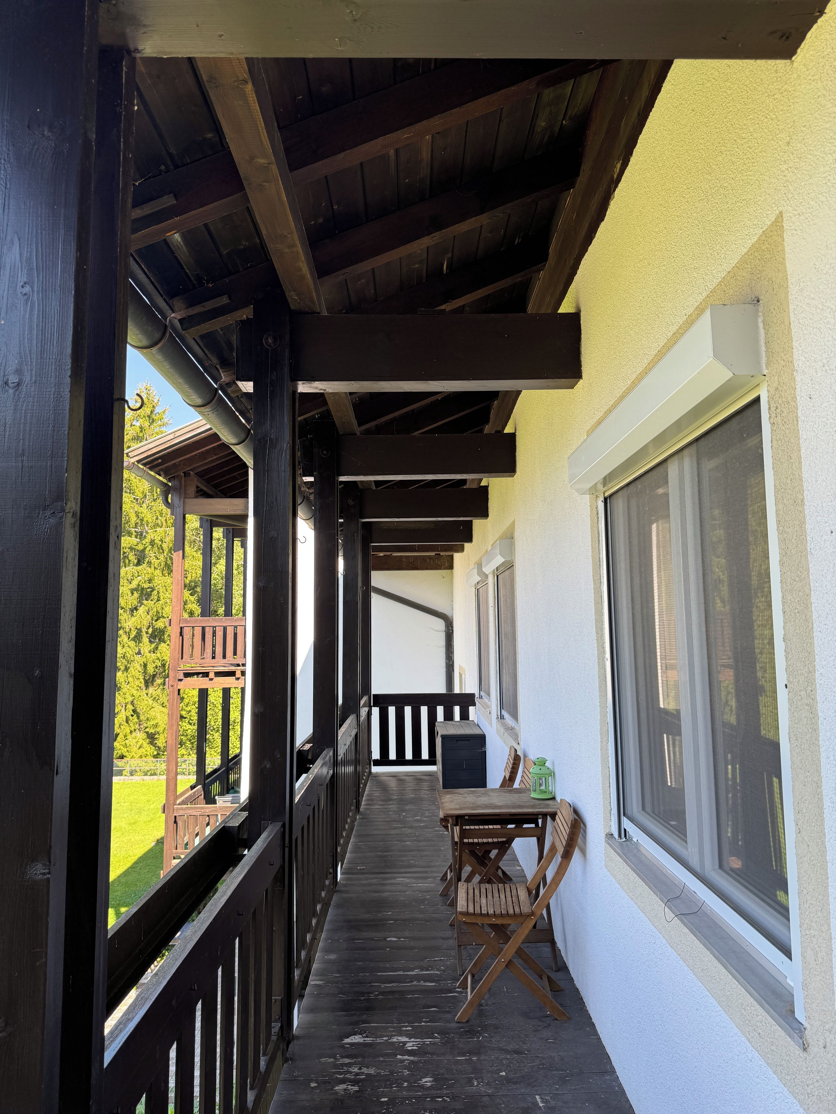 A wooden balcony with a table and chairs, a window, and a view of greenery.