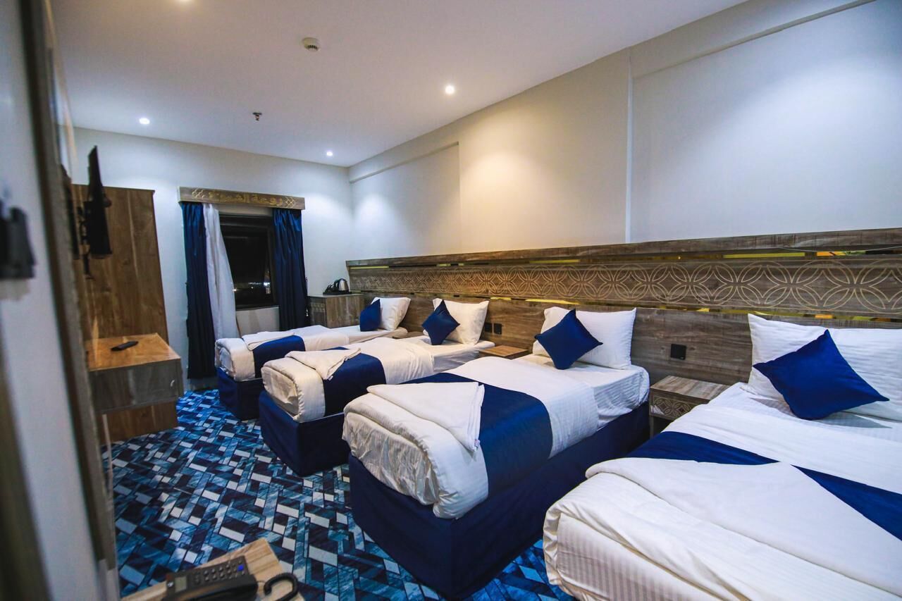 A hotel room with four beds arranged in a row, a wooden headboard, a patterned floor, and a small table with a remote control.