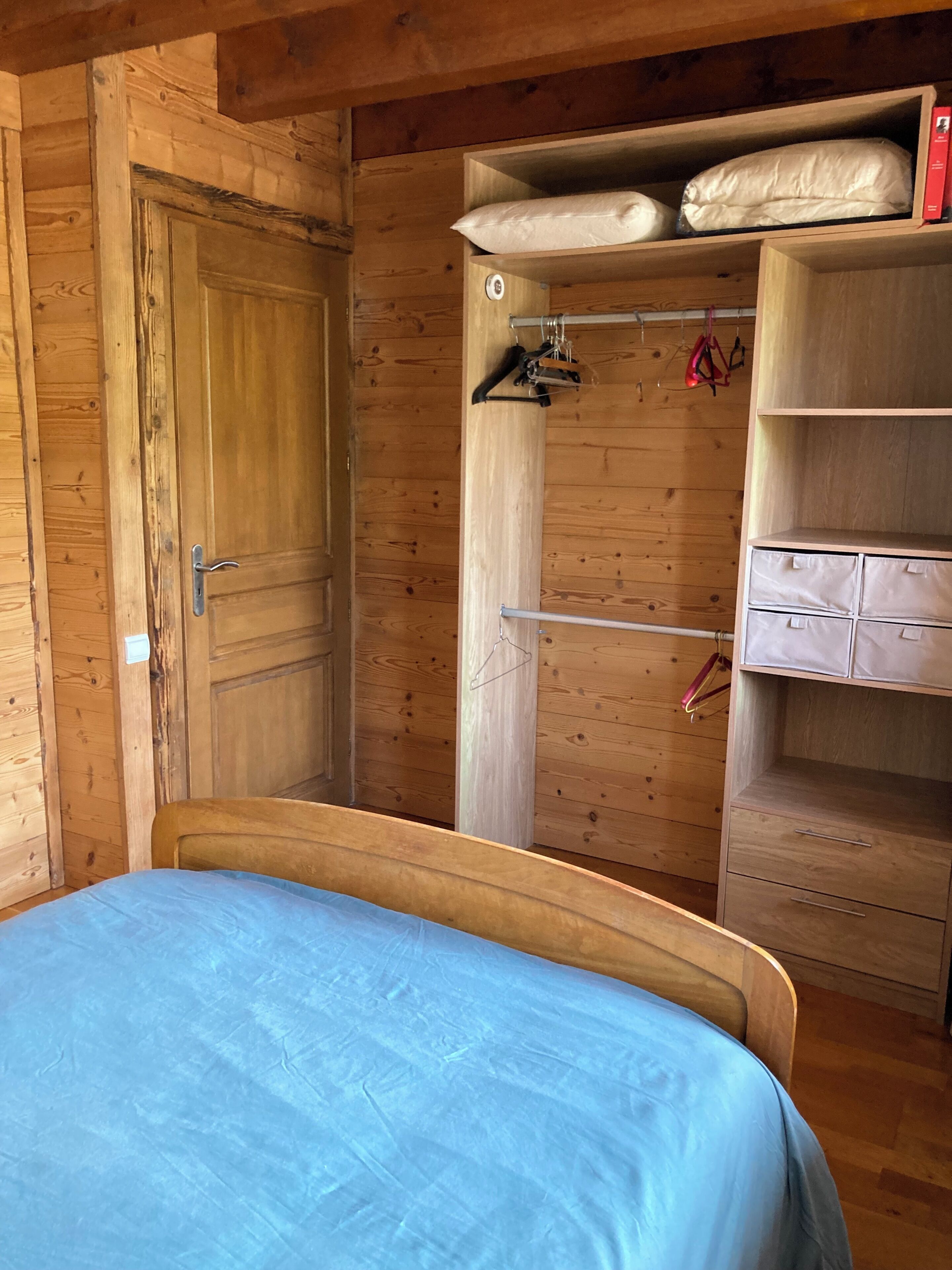 A wooden room with a bed, a wardrobe with hanging space, and a shelf unit.