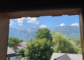 Eine Berglandschaft durch ein Fenster mit Holzrahmen, mit Häusern und Bäumen im Vordergrund.