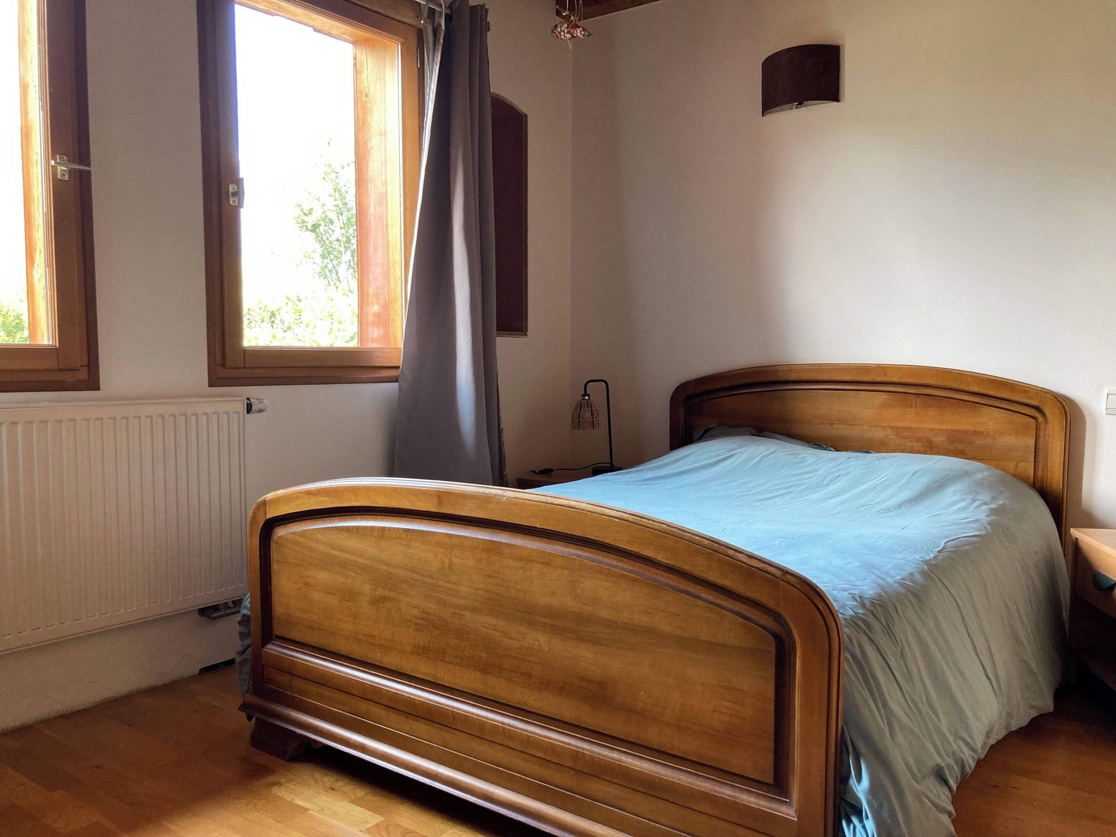 A bedroom with a wooden bed, a radiator, a window with curtains, and a wall-mounted lamp.