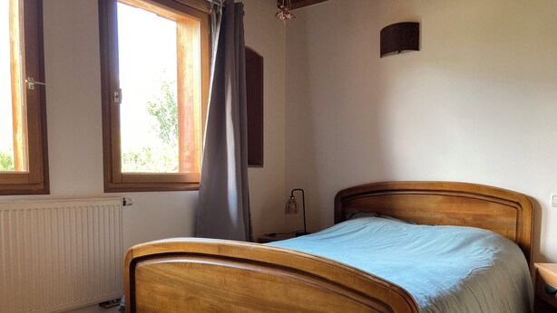 A bedroom with a wooden bed, a radiator, a window with curtains, and a wall-mounted lamp. - Apartment near the lake with a terrace offering mountain views. (Doussard)