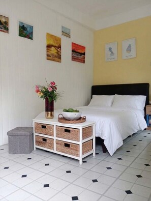 A bedroom with a bed, a bedside table with a vase and a bowl, a small stool, and a wall with framed pictures.