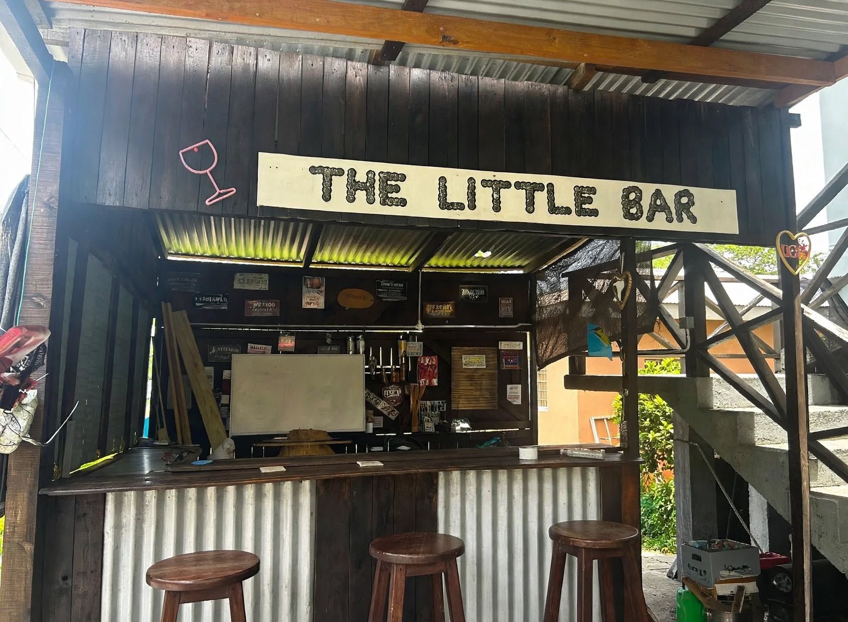 A rustic bar named 'The Little Bar' with a wooden counter, stools, and various signs and decorations.