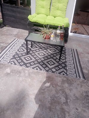Un espace patio agrémenté d’un tapis à motifs, d’une table à plateau en verre, d’un fauteuil vert avec coussin et d’une plante en pot.
