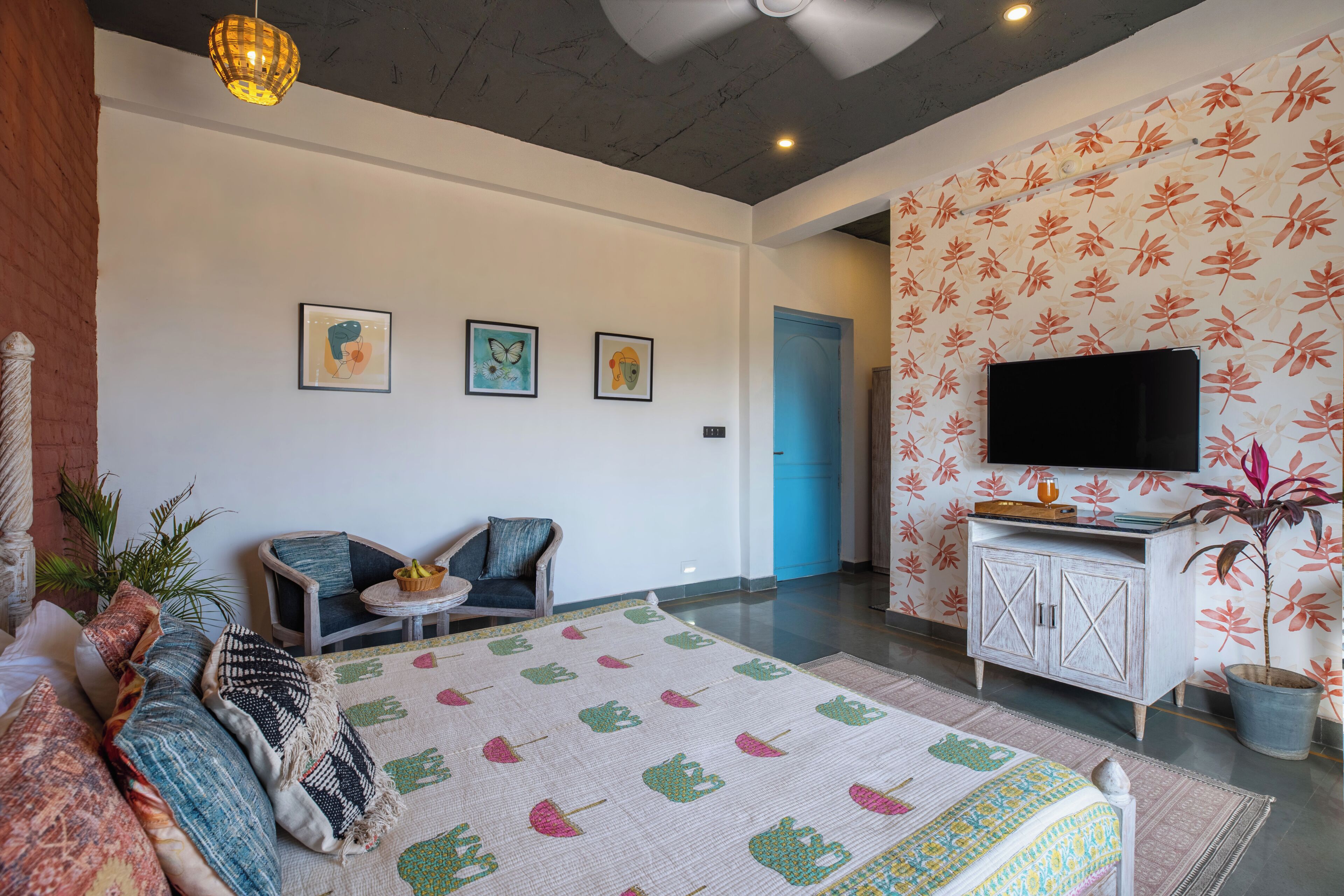 A hotel room with a bed, two chairs, a small table, a TV, and a plant.