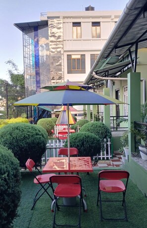 Un espacio al aire libre con mesas y sillas, un paraguas colorido y un edificio al fondo.