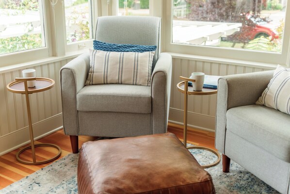 Una habitación acogedora con un sillón de patrón espiga, un reposapiés a juego y un sofá con cojines a rayas. Hay dos mesitas auxiliares con tazas y libros.