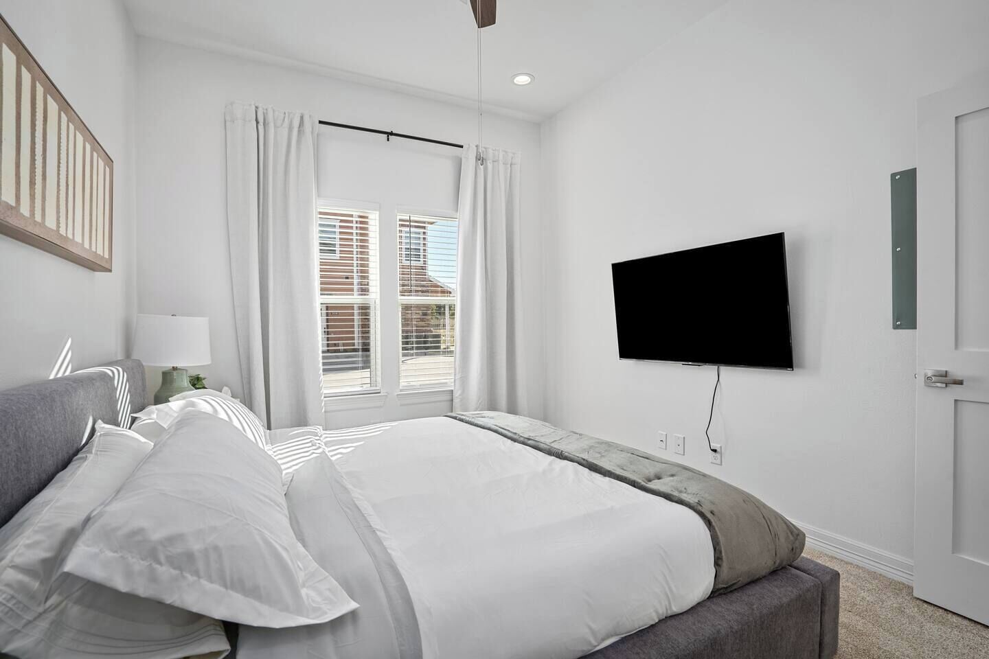 Ein Schlafzimmer mit Bett, einem an der Wand montierten Fernseher, einem Fenster mit Vorhängen und einer Tür.
