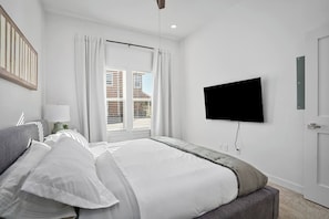 A bedroom with a bed, a TV mounted on the wall, a window with curtains, and a door.