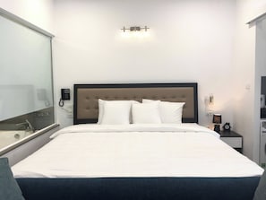 A hotel room with a bed, a bedside table with a lamp, a wall-mounted phone, and a mirror.