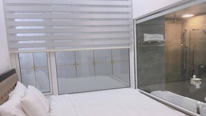 Habitación de hotel con cama, ducha envuelta en vidrio y ventana con persianas.