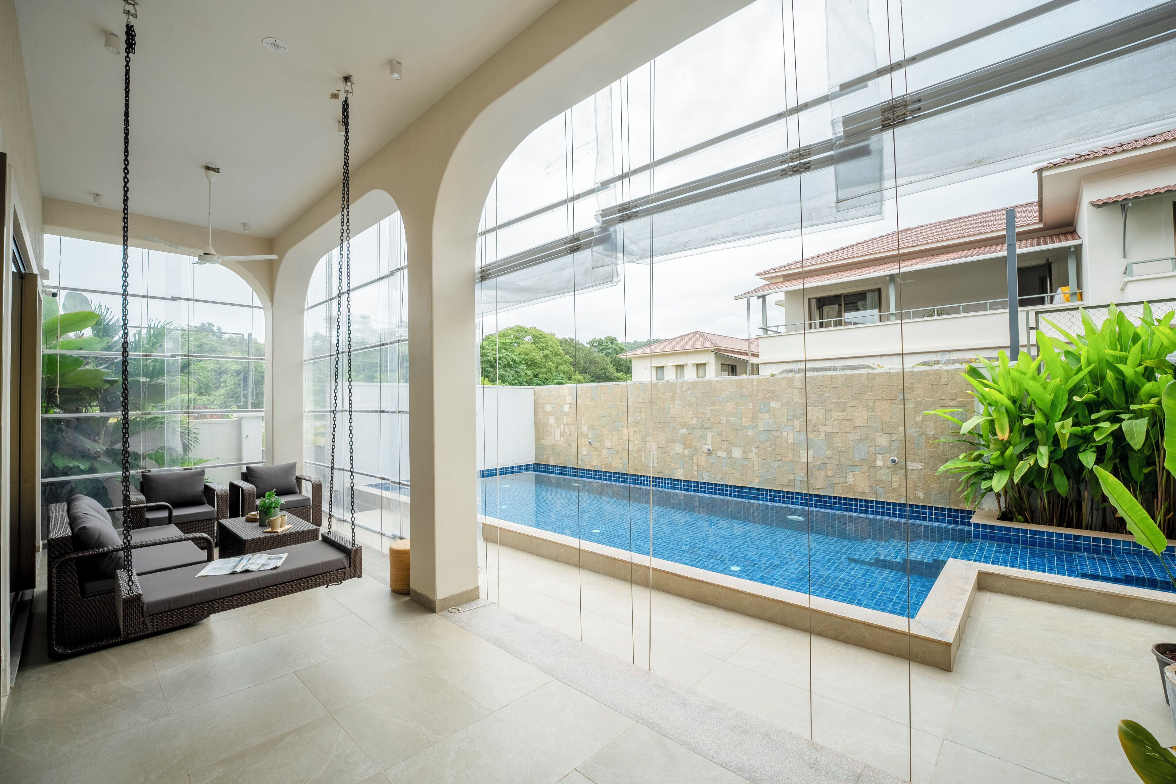 Une piscine intérieure moderne, dotée d'une structure en verre, d'un espace détente avec des meubles en osier et offrant une vue sur un immeuble d'habitation.