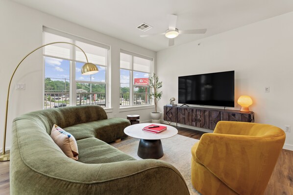 A modern living room with a sofa, armchair, coffee table, TV, and a large window. - 4BDR South Lamar Stay w/ Pool, Rooftop, Restaurant | The Code by AvantStay (Austin)