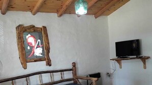 A rustic room with a wooden ceiling, a sofa with a patterned cover, a round table with chairs, a television on a wooden stand, and a wall-mounted picture frame.