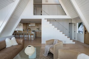 A spacious loft with a high ceiling, a kitchen area, and a staircase leading to an upper level. - A-Frame Cabin w/ Hot Tub + Firepit | EV Charger + Steps to Trails | The A-List (Lake Arrowhead)