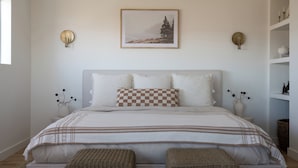 A bedroom with a bed, pillows, a checkered throw, a framed picture, wall sconces, and a shelf with decorative items. - A-Frame Cabin w/ Hot Tub + Firepit | EV Charger + Steps to Trails | The A-List (Lake Arrowhead)