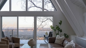 A modern living room with a large glass window, a brown leather sofa, a glass coffee table, and a potted plant. - A-Frame Cabin w/ Hot Tub + Firepit | EV Charger + Steps to Trails | The A-List (Lake Arrowhead)