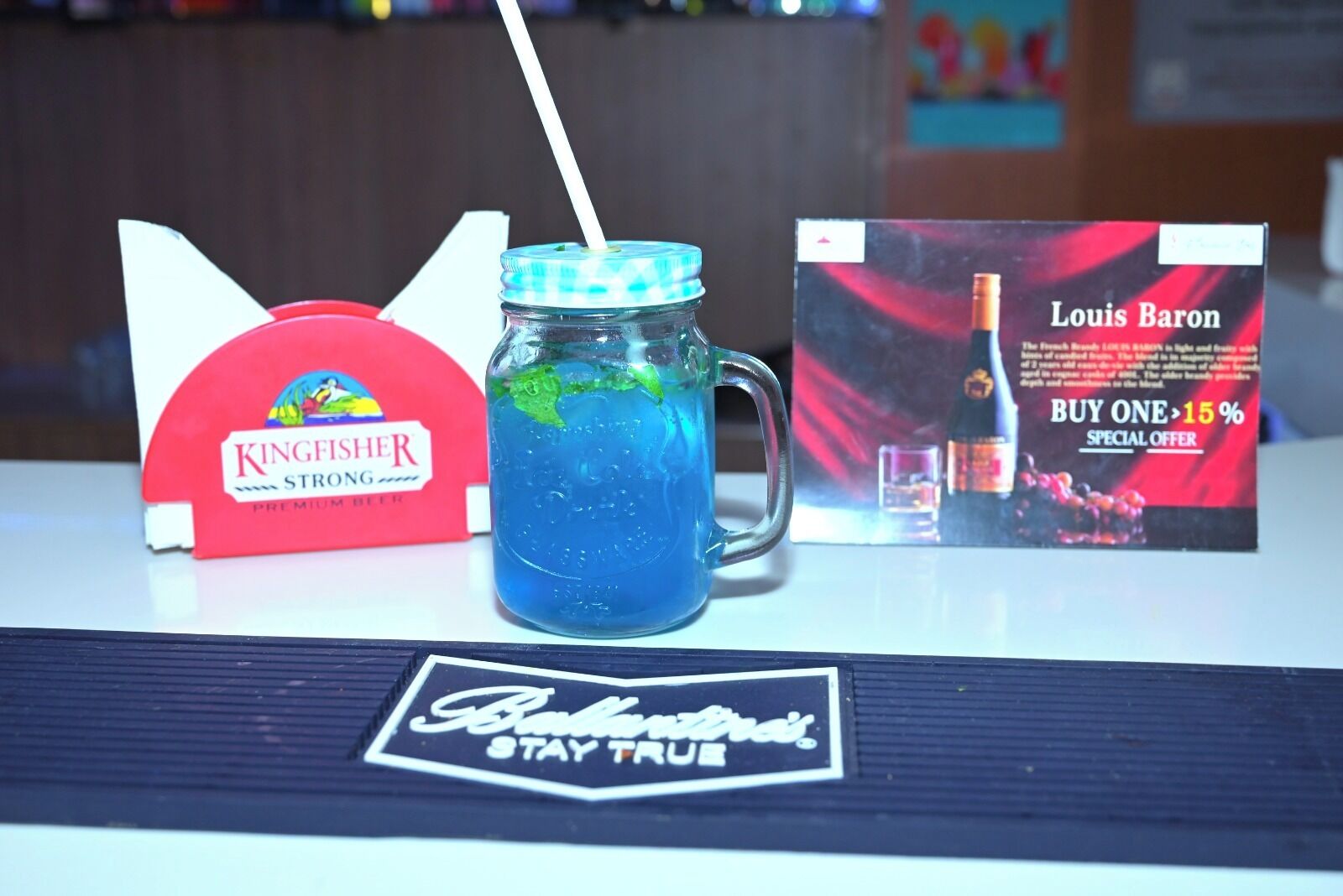 Una caja de cerveza Kingfisher Strong, una bebida azul en un vaso y un folleto promocional de Louis Baron sobre una mesa.
