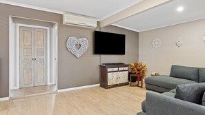 A living room with a flat-screen TV, a grey sofa, a cabinet with decorative items, and a door with a white frame. - Tropical Santai Retreat - Pool with separate flat (Toukley)
