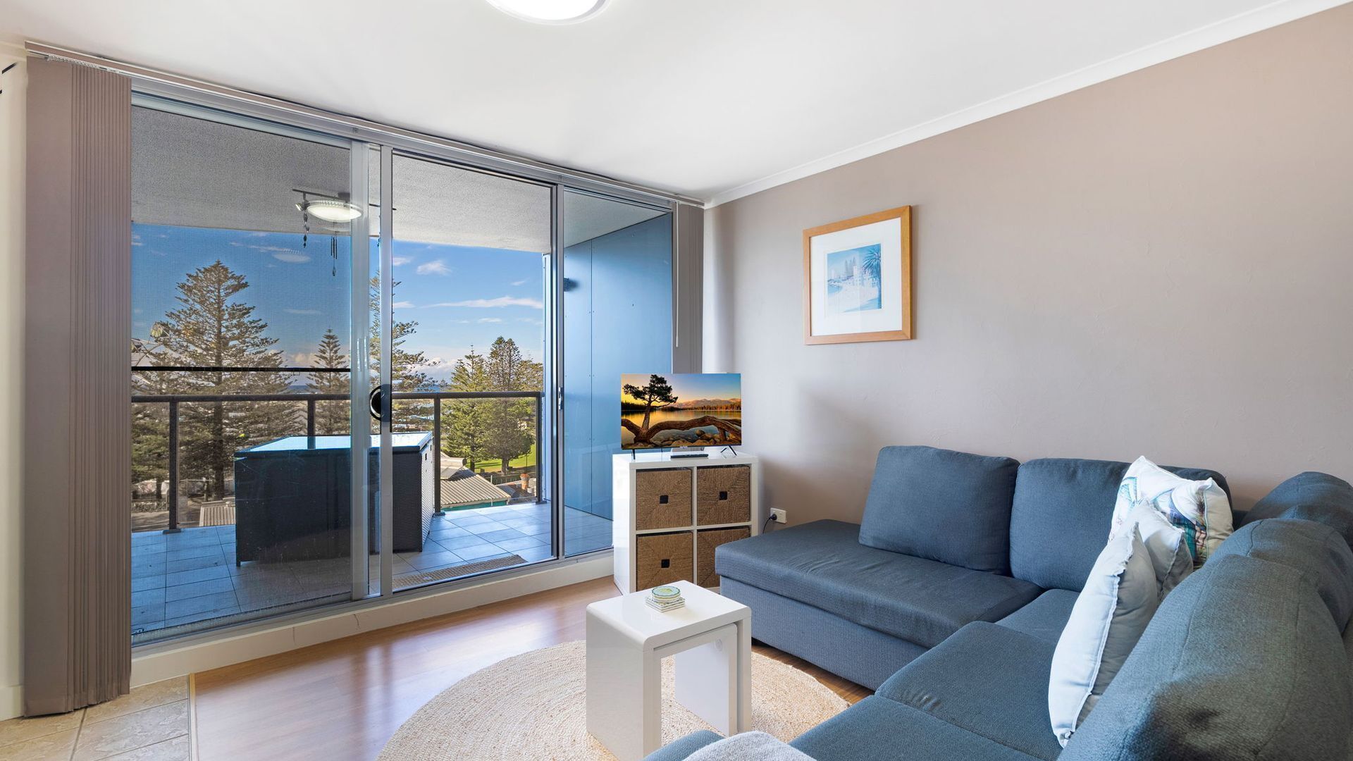 A modern living room with a grey sofa, a small white side table, and a large sliding glass door leading to a balcony with a view of trees and a building.