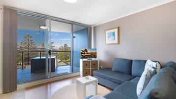 A modern living room with a grey sofa, a small white side table, and a large sliding glass door leading to a balcony with a view of trees and a building.