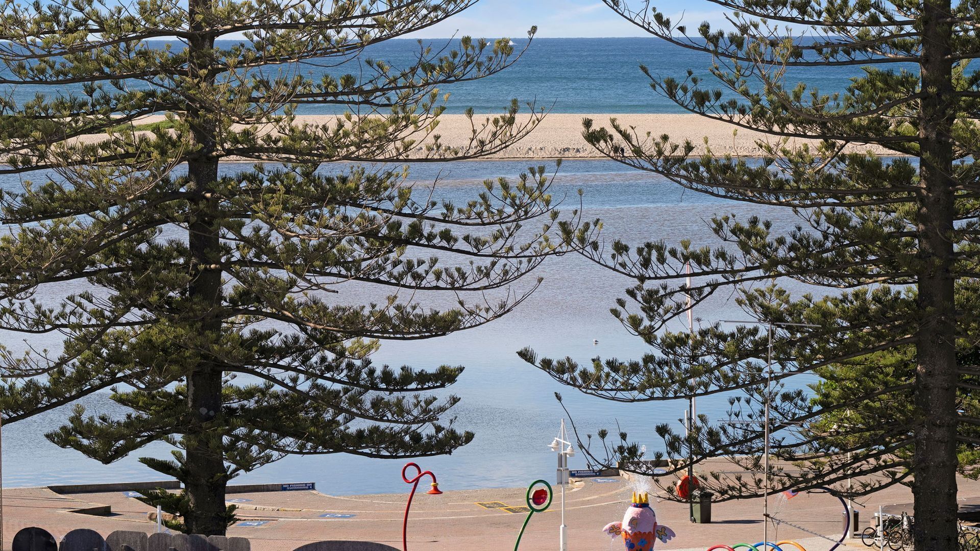 A beach with pine trees in the foreground, a clear sky, and a calm sea.